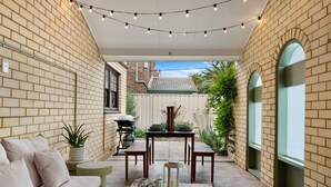 A covered patio with brick walls, a table with chairs, string lights, and a view of a neighboring building. - Sage By The Sea—Stylish Beachside Living with BBQ (West Beach)
