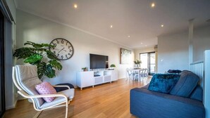 A modern living room with a sofa, a chair, a TV, a clock, and a dining area. - Higher Ground on Seaview - Rooftop Coastal Escape (West Beach)