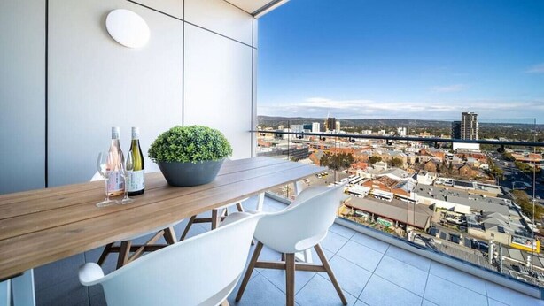 Un balcon moderne avec une table en bois, deux chaises blanches, une plante en pot et deux bouteilles de vin.