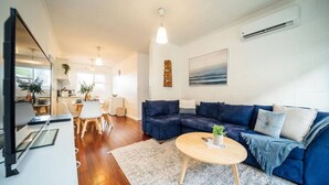 A modern living room with a blue sectional sofa, a round coffee table, and a dining area with a white table and chairs. - Urban Beach Shack - Steps to Grange Beach & Café (Grange)