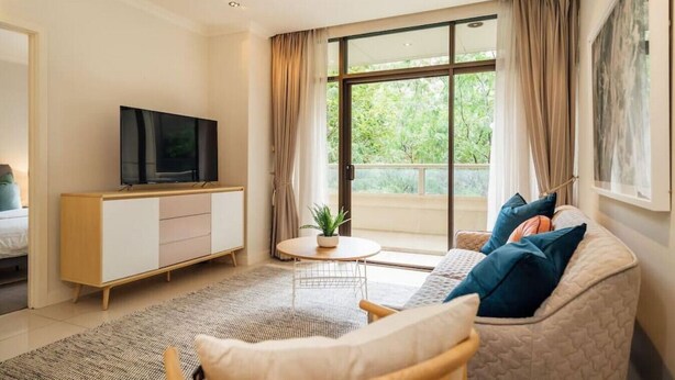 A modern living room with a sofa, TV, and a view of greenery through large windows. - Serene on Grenfell - A Vibrant City Stay (Adelaide)