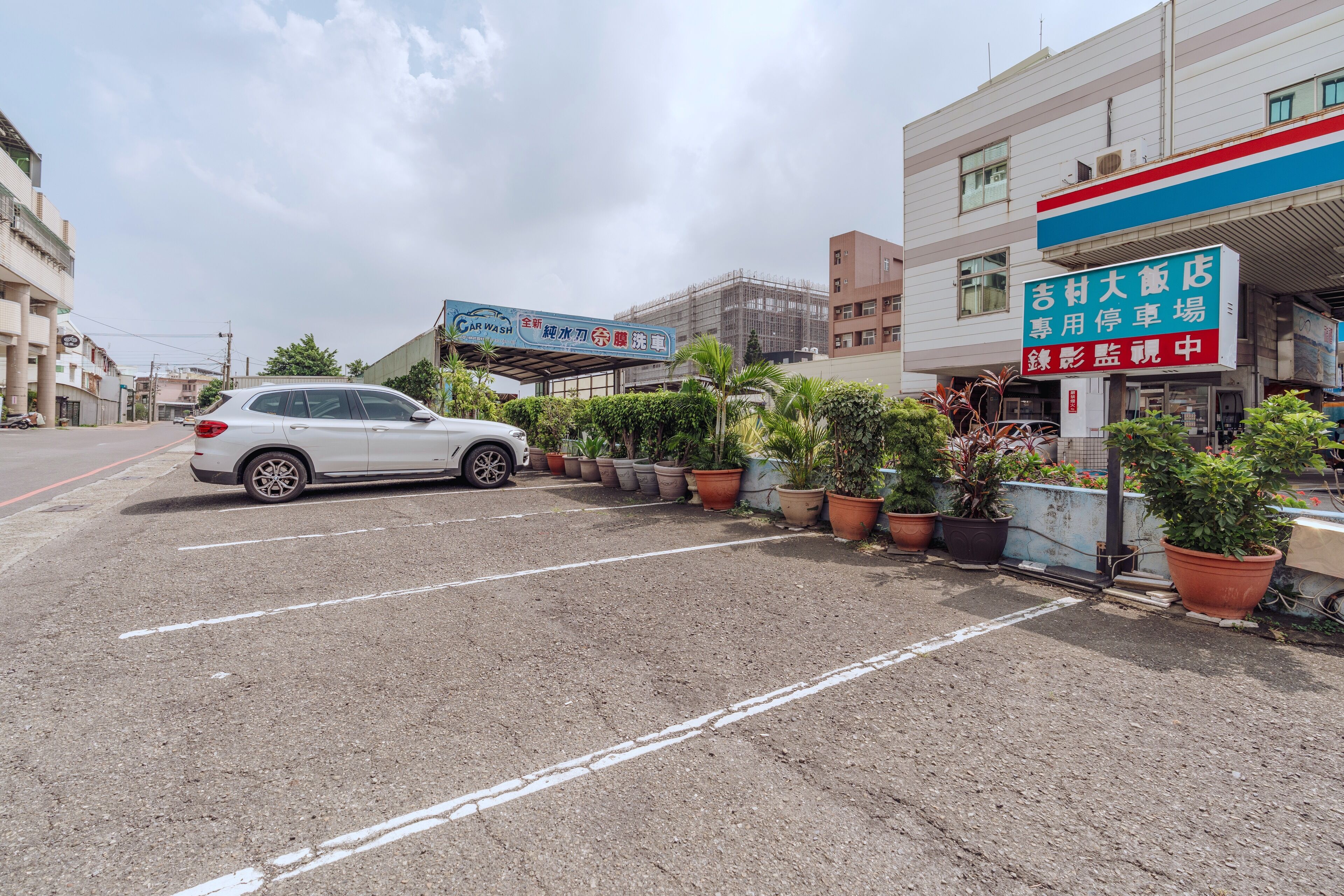 Un estacionamiento con un automóvil blanco, plantas en macetas y un edificio con un letrero.