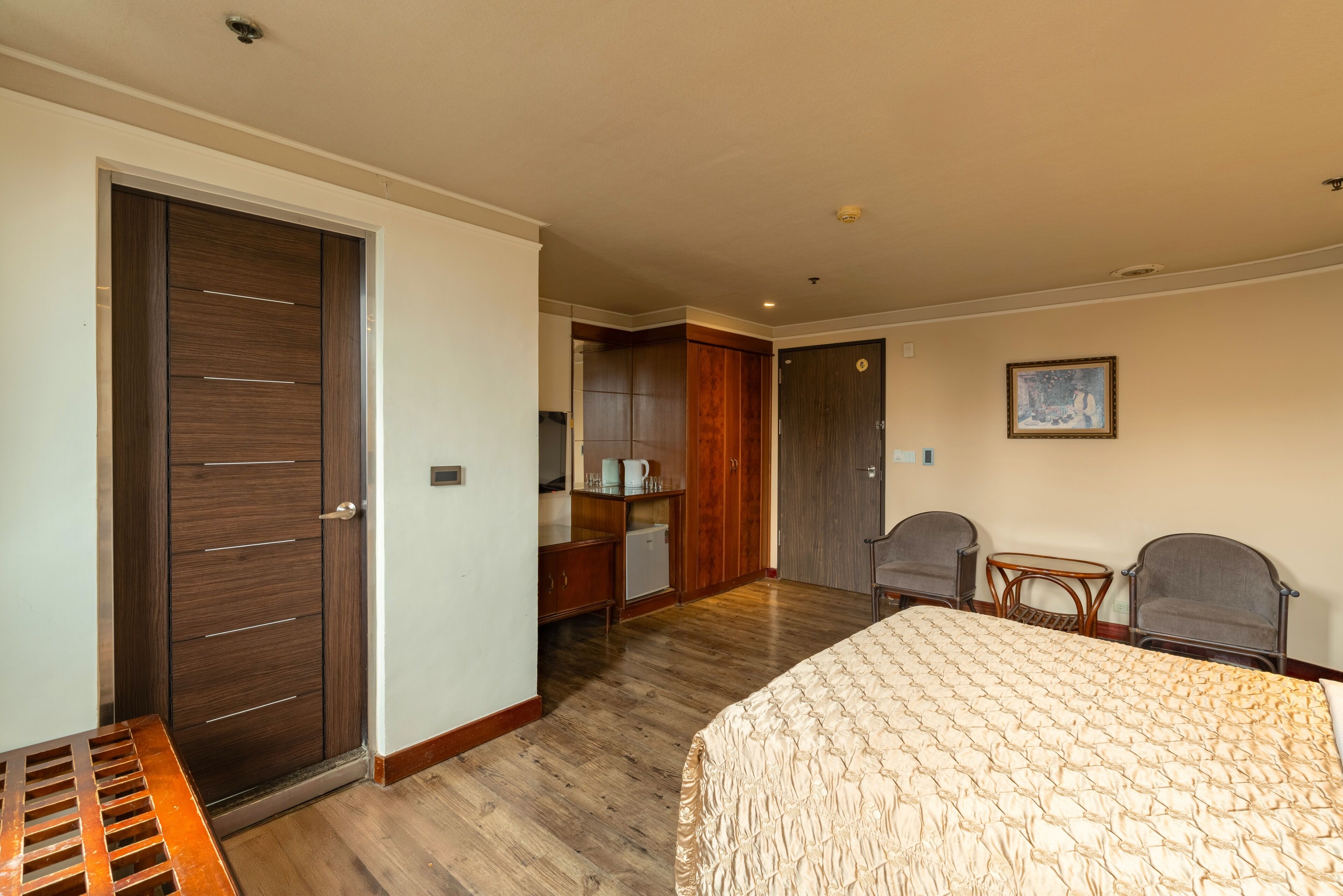 A hotel room with a large bed, a desk, two chairs, a wardrobe, and a small kitchenette.