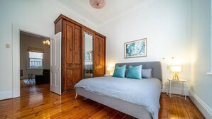 A bedroom with a large bed, wooden wardrobe, and a small bedside table with a lamp. - Irma`s Place—A Historic Garden Cottage (Adelaide)