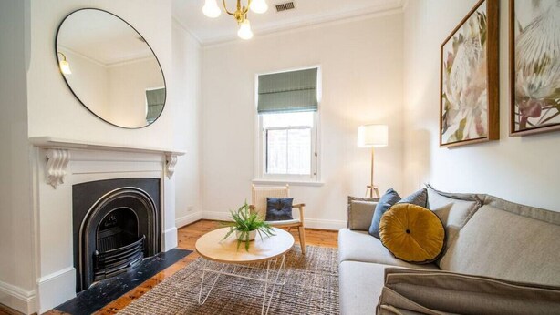 A living room with a fireplace, a round coffee table, a sofa with cushions, a floor lamp, and a large mirror on the wall. - Irma`s Place—A Historic Garden Cottage (Adelaide)