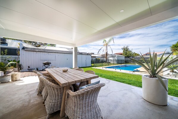 Eine überdachte Terrasse mit einem Holztisch und Korbstühlen, ein Schwimmbecken und der Blick auf umliegende Häuser.