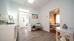 A modern living room with a wooden floor, a white sofa, a chair, and a painting on the wall. - Summer Breeze—A Coastal Poolside Retreat (Glenelg East)