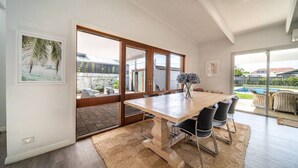 A modern dining area with a large wooden table, black chairs, and a view of a backyard with outdoor furniture. - Summer Breeze—A Coastal Poolside Retreat (Glenelg East)