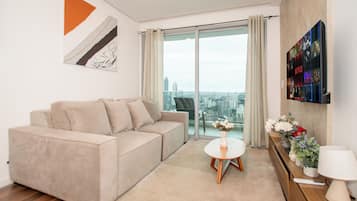 A modern living room with a beige sofa, a small white table with a vase, and a city view through large glass doors.