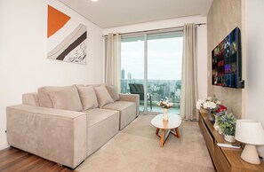 A modern living room with a beige sofa, a small white table with a vase, and a city view through large glass doors.