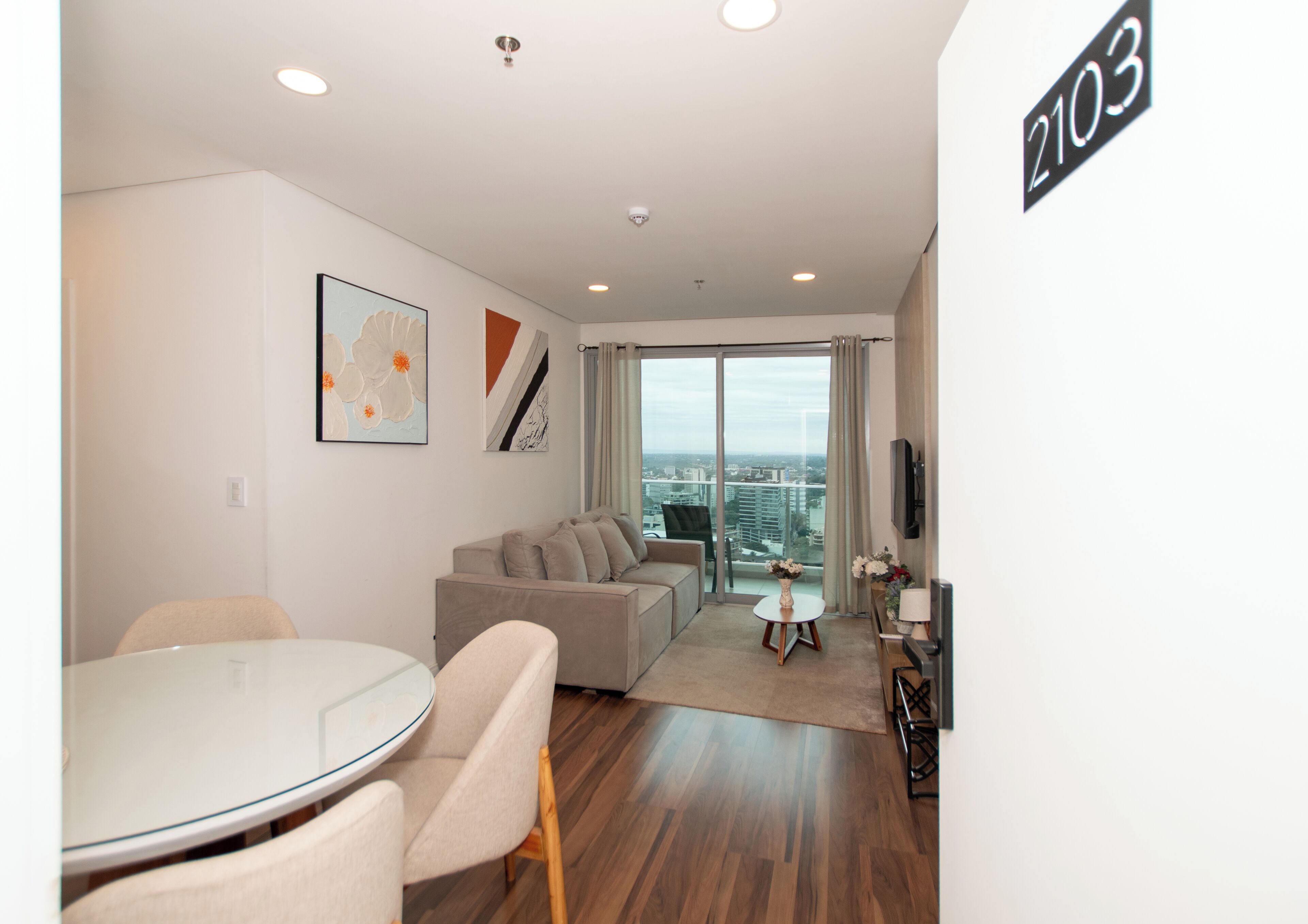 Ein modernes Wohnzimmer mit Glastür zum Balkon, einem Essbereich mit rundem Tisch und einem an der Wand befestigten Flachbildfernseher.