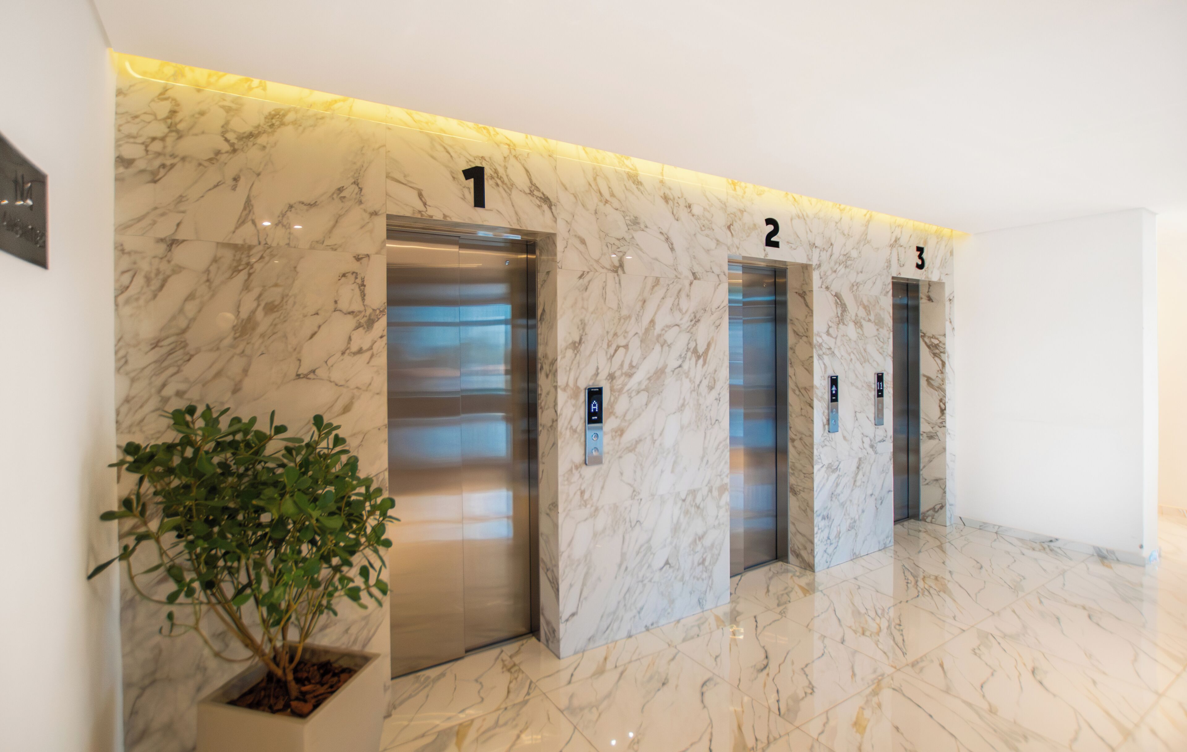 Un vestíbulo de ascensor moderno con paredes y suelos de mármol, números del 1 al 3, y una planta en maceta.