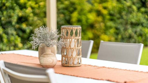 A table with a woven candle holder and a vase with dried plants, set on a tablecloth.