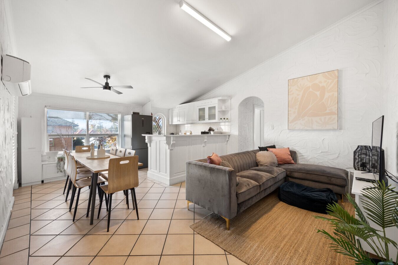 Un vaste espace de vie comprenant une table à manger, un canapé gris et une cuisine à l’arrière-plan.