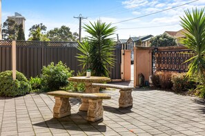 A courtyard with a stone table and benches, surrounded by a fence and various plants.