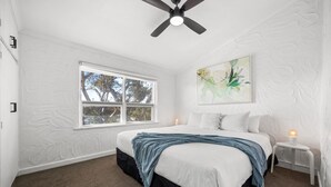 Une chambre avec un grand lit, un ventilateur de plafond, une fenêtre donnant sur les arbres et un tableau accroché au mur.
