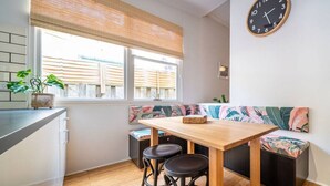 Una cocina moderna con una mesa de madera, dos taburetes, un banco con cojín estampado tropical, un gran reloj de pared y una ventana con persianas.