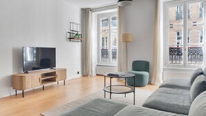 A modern living room with a flat-screen TV, a wooden cabinet, a round coffee table, a teal armchair, and a grey sofa.