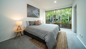 A bedroom with a large bed, a bedside table with a lamp, a painting on the wall, and a large window with a view of trees. - Juniper on Rundle - A Leafy City-fringe Escape (Kent Town)