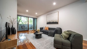 A modern living room with a grey sofa, a coffee table, and a TV on a wooden stand. - Juniper on Rundle - A Leafy City-fringe Escape (Kent Town)