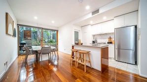 A modern kitchen with a large island, wooden flooring, and a view of a garden through glass doors. - Juniper on Rundle - A Leafy City-fringe Escape (Kent Town)