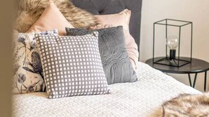 A neatly made bed with a grey headboard, multiple pillows, and a decorative lamp on a small side table.
