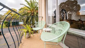 A balcony with two green chairs, a potted plant, and a view of a tree and a building across the street.