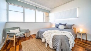 A bedroom with a large window, a bed with grey bedding, a grey armchair, a small side table with a lamp, and a woven rug.