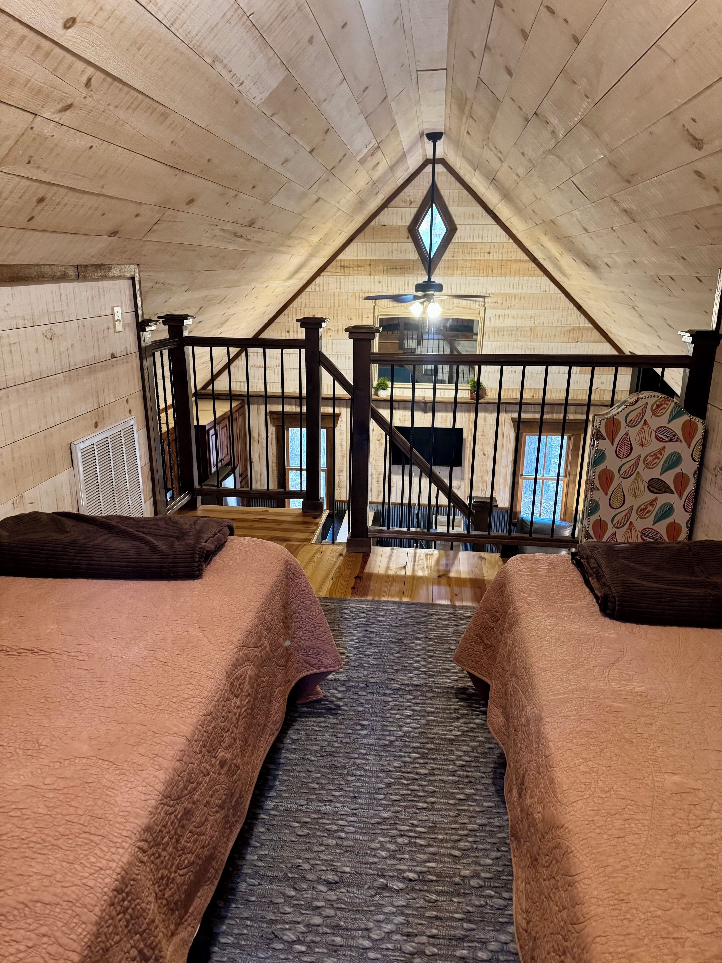 A cozy attic bedroom with two beds, a wooden ceiling, a staircase, and a view of the outside.