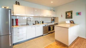 A modern kitchen with white cabinets, a stainless steel refrigerator, a dishwasher, and a small island with a sink. - Get Comfy on Frew - CBD Stay near Parklands & Dining (Adelaide)