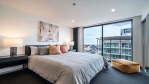 A modern hotel room with a large bed, two bedside tables with lamps, a large window with a city view, and a painting on the wall.