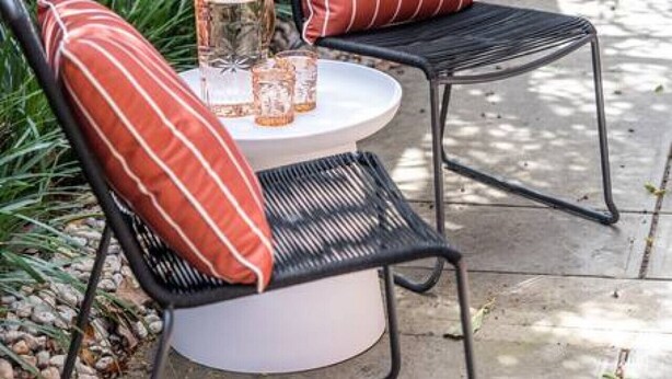 Deux chaises en métal noir avec des coussins à rayures rouges, une table ronde blanche avec un porte-bougie en verre et un petit vase, le tout placé sur une terrasse en béton, avec une clôture en métal et de la verdure en arrière-plan.