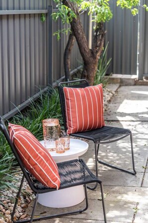 Deux chaises en métal noir avec des coussins à rayures rouges, une table ronde blanche avec un porte-bougie en verre et un petit vase, le tout placé sur une terrasse en béton, avec une clôture en métal et de la verdure en arrière-plan.