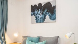 Un lit bien fait, avec une couette de couleur claire et des coussins turquoise, une peinture abstraite moderne accrochée au mur et une lampe de chevet posée sur une table de nuit en bois.