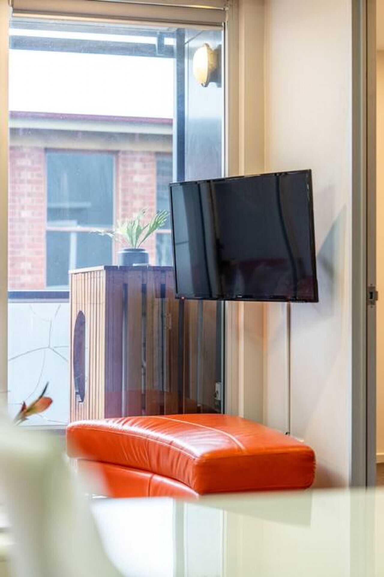 Una habitación moderna con una televisión de pantalla plana montada sobre un soporte, una planta en maceta y un banco acolchado de color naranja brillante.