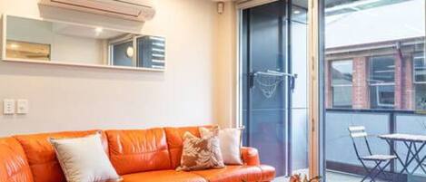 A modern living room with a large orange sectional sofa, a white coffee table, and a glass door leading to an outdoor area with a table and chairs.