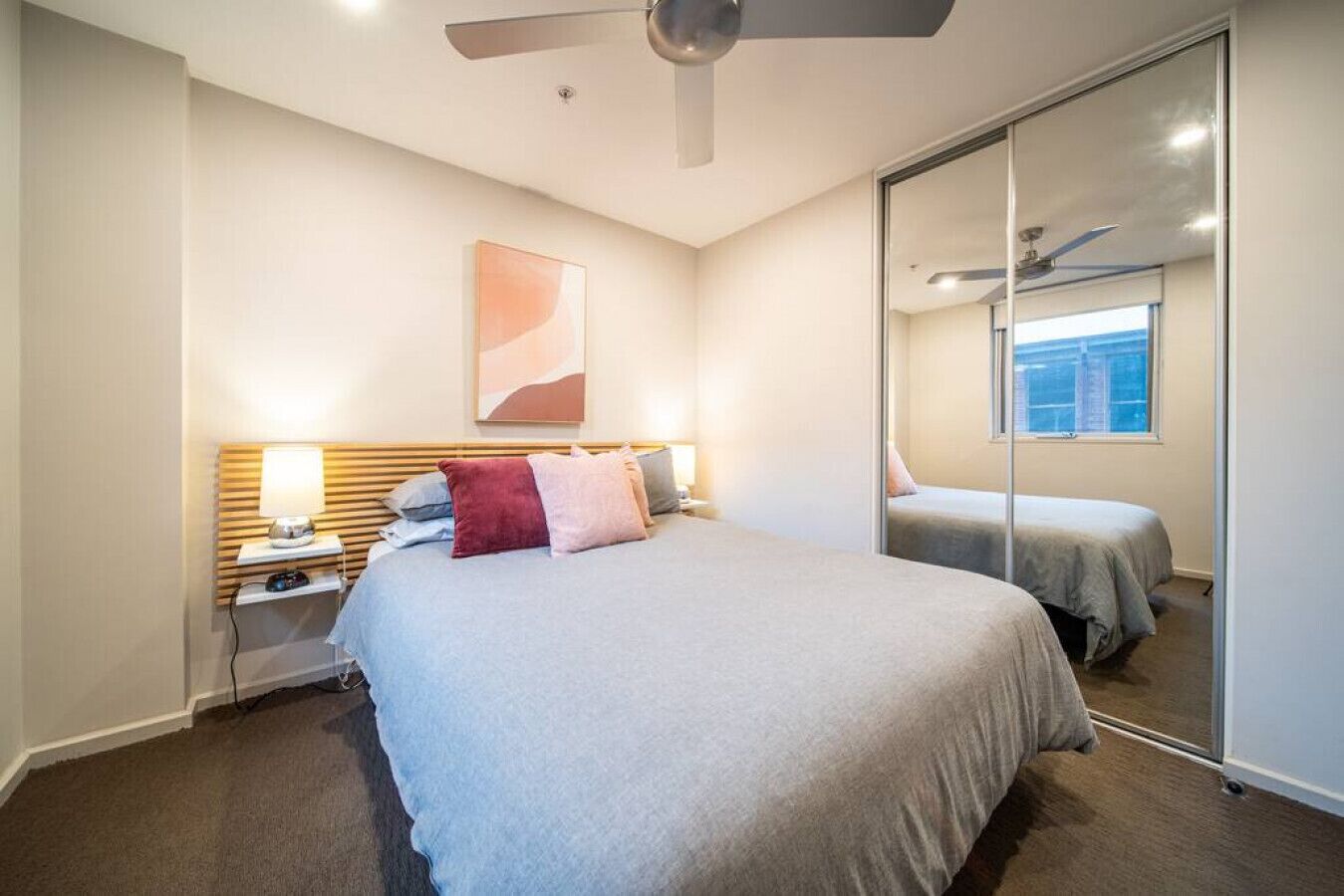 Une chambre d’hôtel moderne avec un grand lit, des lampes de chevet, un ventilateur de plafond et un grand miroir qui reflète la pièce.