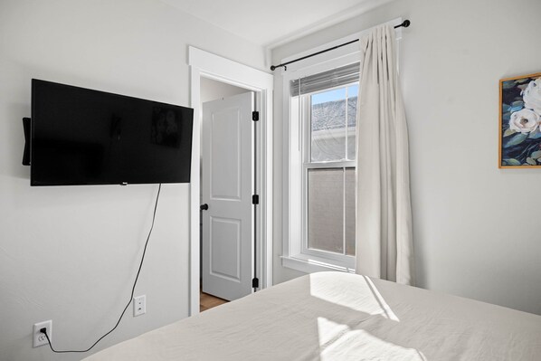 Une chambre à coucher avec un lit, un téléviseur fixé au mur, une fenêtre avec des rideaux et un tableau accroché au mur.