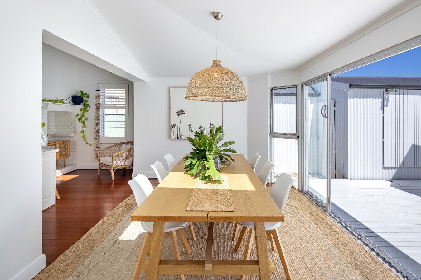 A dining area with a long wooden table, white chairs, a woven pendant light, and a large window with a view.