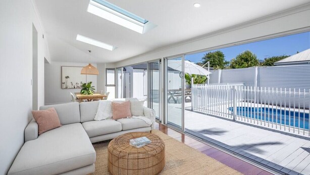 A modern living room with a large sectional sofa, a round wicker coffee table, and a view of a swimming pool through large glass doors. - Villa 1 @ THE OSLO on Broadway with Pool (Glenelg South)