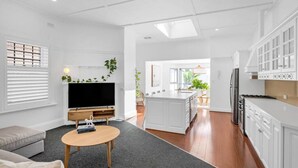 A modern living room with a flat-screen TV, a coffee table, and a sofa. The kitchen features white cabinets and a large island. - Villa 1 @ THE OSLO on Broadway with Pool (Glenelg South)