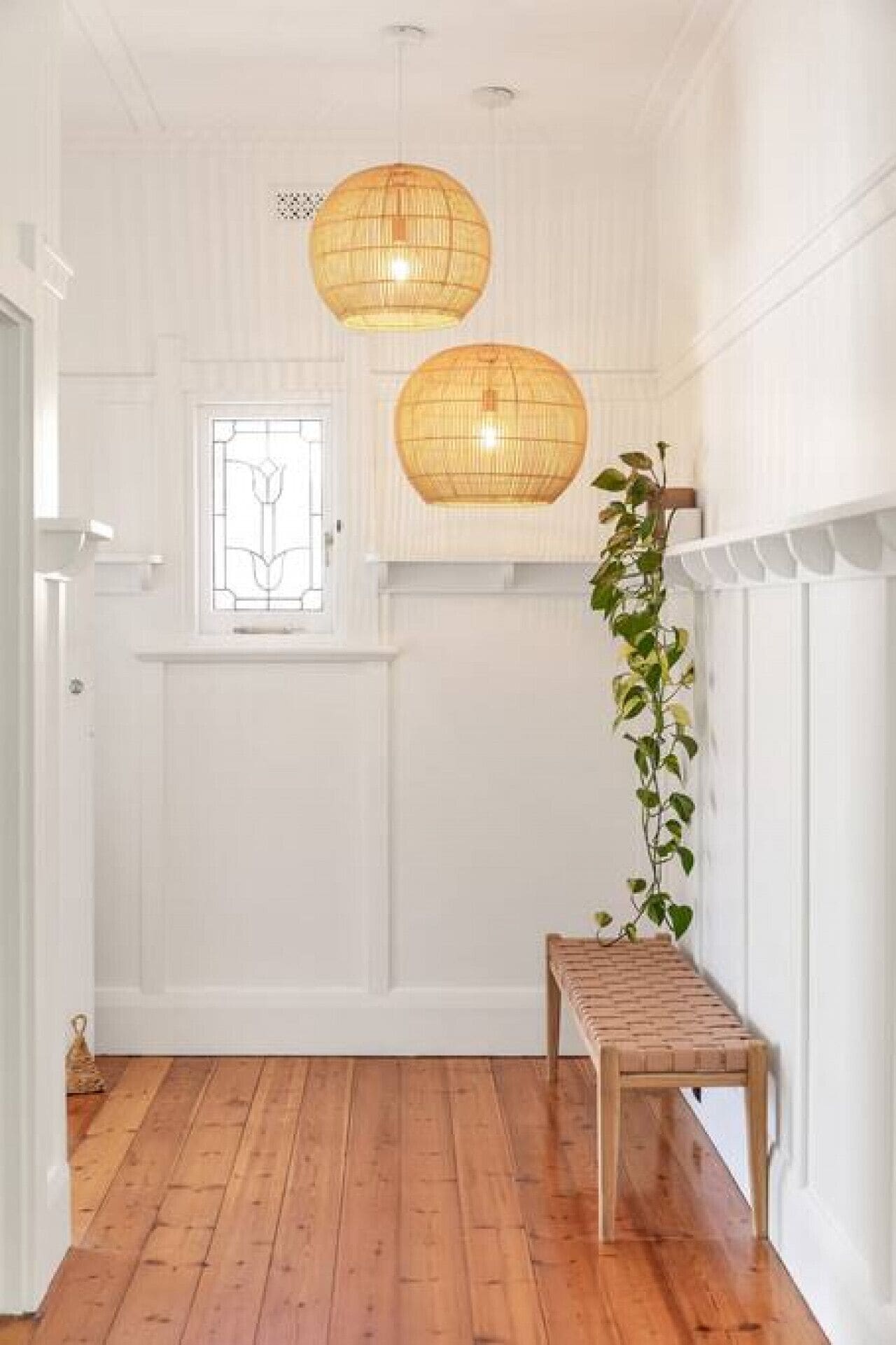 A hallway with wooden flooring, white walls, a wooden bench, and two hanging wicker lamps.