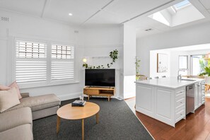 A modern living room with a sofa, TV, and a kitchen area with a white cabinet and sink. - THE OSLO on Broadway—Two Poolside Villas (Glenelg South)