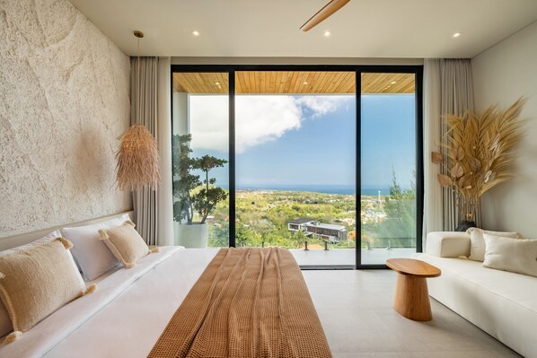 A modern bedroom with a large bed, a wooden side table, a sofa, and a large window offering a view of the landscape.