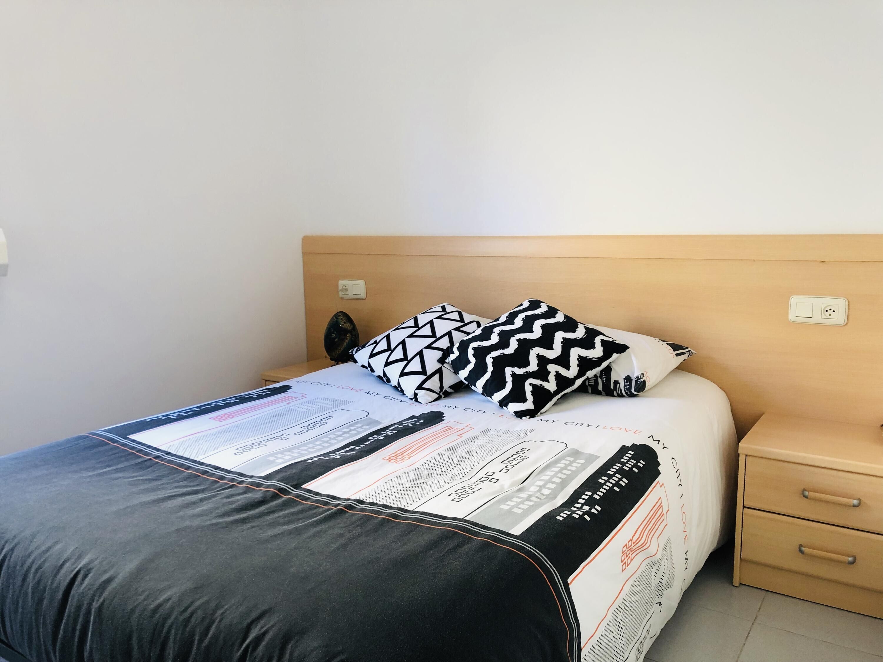 Uma cama bem arrumada com almofada estampada em preto e branco, cabeceira de madeira e mesa de cabeceira com gaveta.