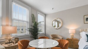 Una habitación con una mesa redonda, sillas anaranjadas, una cama, una mesita de noche, una lámpara, un espejo y un ventanal grande con persianas.