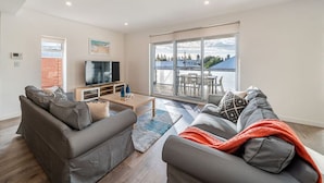 A modern living room with a grey sofa set, a coffee table, and a TV. There is a balcony with a view of the sky and buildings. - Level Up Henley - Modern Comfort near Beach & Dining (Henley Beach)