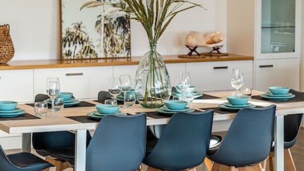 A modern dining area with a long table set for six, featuring teal and white tableware, black chairs, and a glass vase with plants. - Level Up Henley - Modern Comfort near Beach & Dining (Henley Beach)