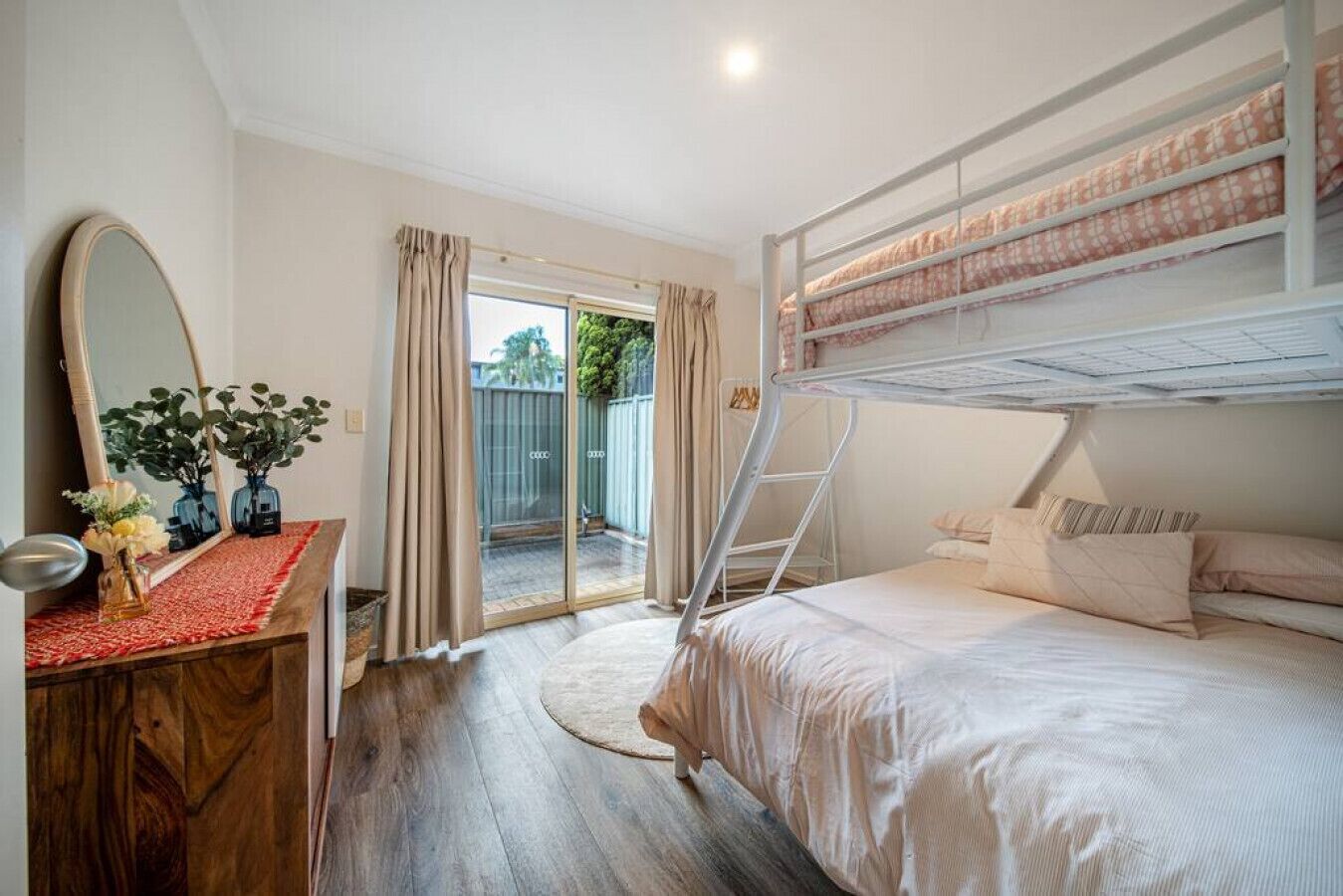 Een slaapkamer met een stapelbed, een houten dressoir, een spiegel en een schuifdeur naar een buitenruimte.