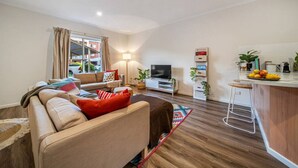 Una sala de estar moderna con un sofá, televisión y una pequeña zona de cocina con un taburete y fruta fresca.